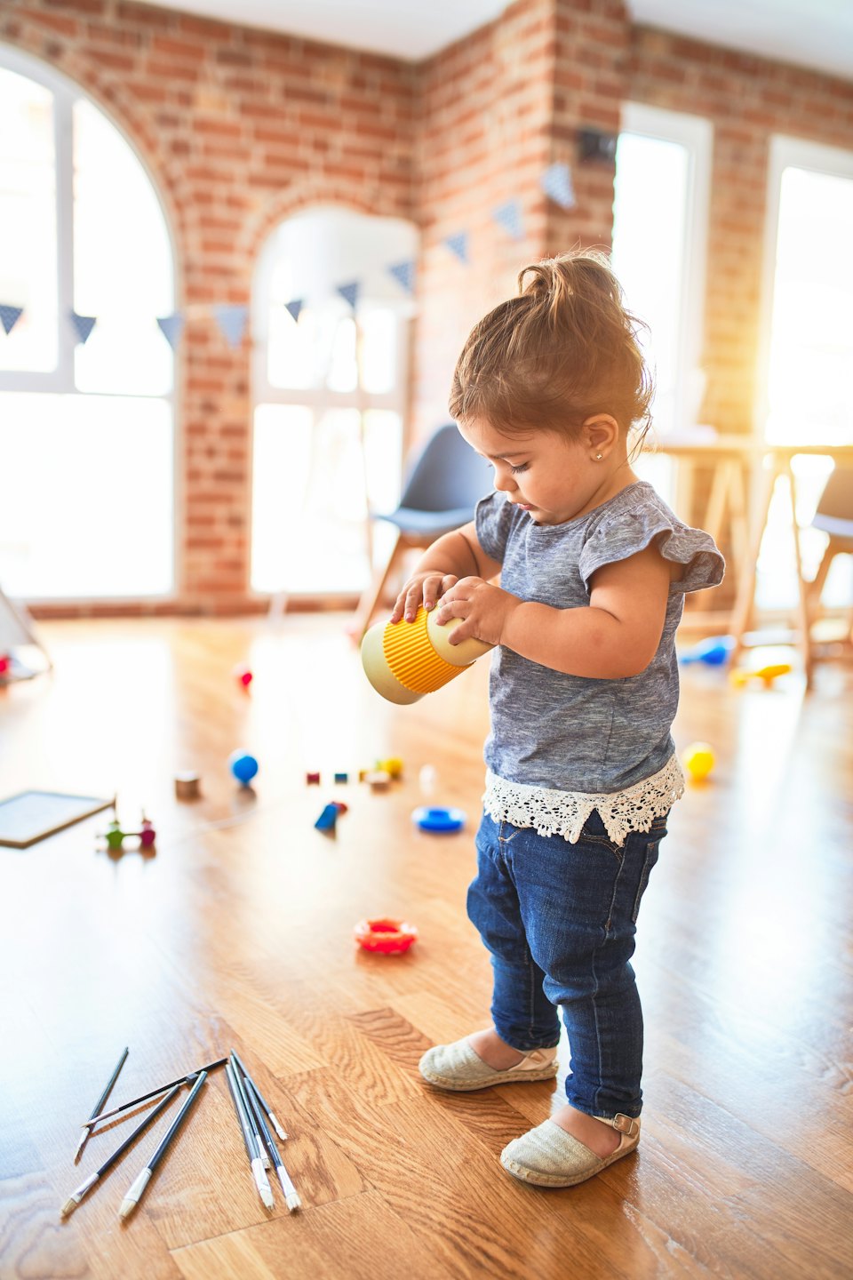 Toddler plays with toy cup on wooden floor near brick wall.